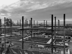 A construction site with an unfinished steel framework structure, surrounded by trees and a cloudy sky. Various construction materials and vehicles are visible at ground level within the site.