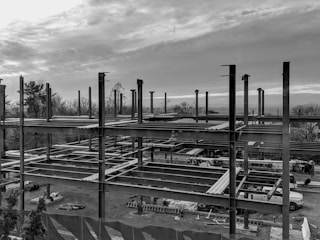 A construction site with an unfinished steel framework structure, surrounded by trees and a cloudy sky. Various construction materials and vehicles are visible at ground level within the site.