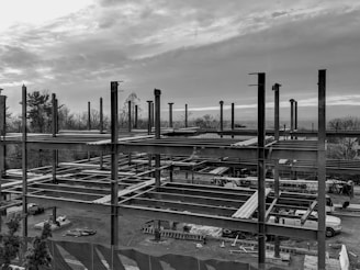 A construction site with an unfinished steel framework structure, surrounded by trees and a cloudy sky. Various construction materials and vehicles are visible at ground level within the site.