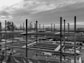 A construction site with an unfinished steel framework structure, surrounded by trees and a cloudy sky. Various construction materials and vehicles are visible at ground level within the site.