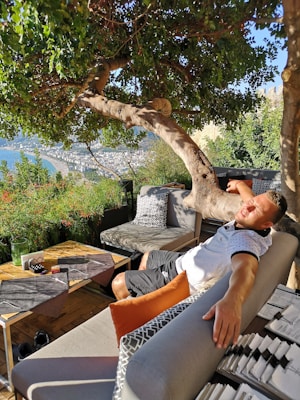 A person lounging on a comfortable outdoor sofa under a large, leafy tree. The setting offers a scenic view of a city and the ocean. The area is furnished with cushions, a wooden table with place settings, and is surrounded by lush greenery.