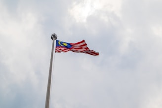 A Malaysian flag is waving on a tall flagpole against a backdrop of overcast skies. The flag features red and white stripes with a blue canton containing a yellow crescent and star.