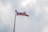 A Malaysian flag is waving on a tall flagpole against a backdrop of overcast skies. The flag features red and white stripes with a blue canton containing a yellow crescent and star.