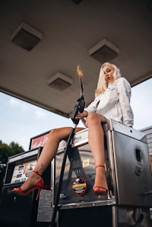 A woman with blonde hair sits on top of a vintage fuel pump wearing a white coat and red high heels. She holds a fuel nozzle with flames coming out of the end, creating a striking and edgy pose. The setting appears to be an old gas station.