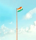 A tall flagpole with the Indian national flag waving at the top against a clear, bright sky. The sky features a gradient of blue hues with scattered white clouds.