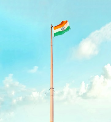 A tall flagpole with the Indian national flag waving at the top against a clear, bright sky. The sky features a gradient of blue hues with scattered white clouds.