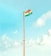 A tall flagpole with the Indian national flag waving at the top against a clear, bright sky. The sky features a gradient of blue hues with scattered white clouds.