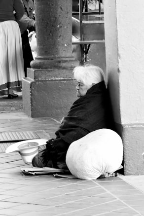 An elderly person with white hair sits on the ground, leaning against a wall. They are wrapped in a dark blanket. A large cloth sack is placed beside them, and a hat is resting on the ground nearby. The scene is monochromatic, with various shades of gray. Other people and architectural elements are partially visible in the background.