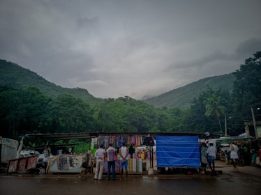 A small marketplace situated in a mountainous area with lush greenery. Several people are gathered around various stalls that sell fabrics and other goods. The sky is overcast, suggesting an impending rain. The surrounding hills are covered with dense forest.