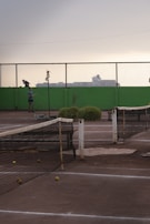 A tennis court with a worn-out net and several tennis balls scattered on the ground. In the background, there is a person standing next to the court with a tennis racket. The scenery includes a high green wall and a mesh fence, with industrial buildings silhouetted against an overcast sky.
