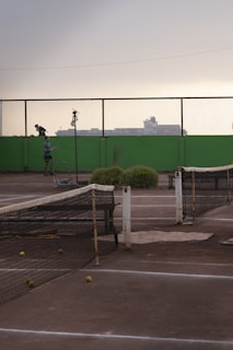 A tennis court with a worn-out net and several tennis balls scattered on the ground. In the background, there is a person standing next to the court with a tennis racket. The scenery includes a high green wall and a mesh fence, with industrial buildings silhouetted against an overcast sky.