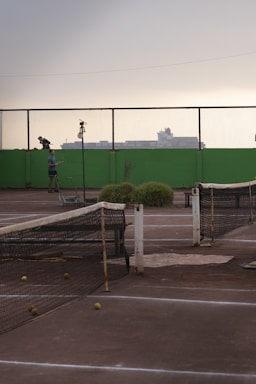 A tennis court with a worn-out net and several tennis balls scattered on the ground. In the background, there is a person standing next to the court with a tennis racket. The scenery includes a high green wall and a mesh fence, with industrial buildings silhouetted against an overcast sky.