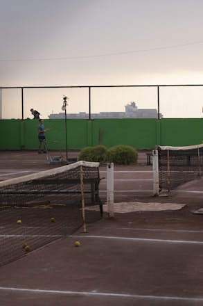 A tennis court with a worn-out net and several tennis balls scattered on the ground. In the background, there is a person standing next to the court with a tennis racket. The scenery includes a high green wall and a mesh fence, with industrial buildings silhouetted against an overcast sky.