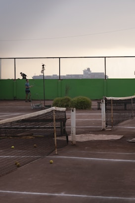 A tennis court with a worn-out net and several tennis balls scattered on the ground. In the background, there is a person standing next to the court with a tennis racket. The scenery includes a high green wall and a mesh fence, with industrial buildings silhouetted against an overcast sky.