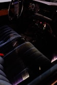 The interior of a car featuring a vintage dashboard with analog gauges and a steering wheel. The seating is dark-colored and plush, possibly made of velvet or a similar fabric. There are controls and a radio on the center console, illuminated by subtle lighting.