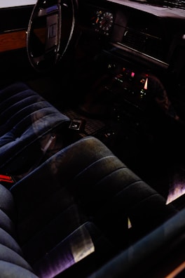 The interior of a car featuring a vintage dashboard with analog gauges and a steering wheel. The seating is dark-colored and plush, possibly made of velvet or a similar fabric. There are controls and a radio on the center console, illuminated by subtle lighting.