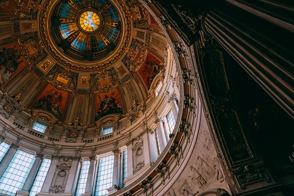 The image shows a view from below, looking up at the dome and columns of a large, ornate building with intricate carvings and paintings on its walls. The dome is painted white and features a stained glass window that casts colorful light throughout the space. The columns are made of marble and stand tall against the wall, adding to the grandeur of the structure. The perspective from below emphasizes the height and scale of the architectural marvel, creating an awe-inspiring scene for viewers.