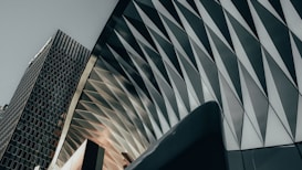A modern architectural structure with a geometric pattern is juxtaposed against a tall, rectangular office building. The pattern on the structure features triangular shapes that create a dynamic, textured surface. The lighting casts subtle shadows that enhance the three-dimensional effect of the design.
