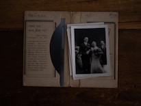 A black and white vintage photograph of a wedding scene is placed on top of an old paper or package. The image includes a bride in a long gown holding a bouquet, standing next to a groom in a suit. Several other figures, possibly wedding guests, are present. The background shows an advertisement for photographic film, suggesting nostalgia and historical context.