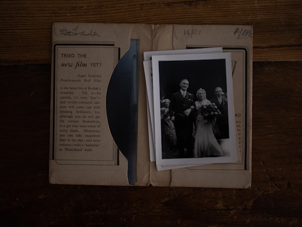 A black and white vintage photograph of a wedding scene is placed on top of an old paper or package. The image includes a bride in a long gown holding a bouquet, standing next to a groom in a suit. Several other figures, possibly wedding guests, are present. The background shows an advertisement for photographic film, suggesting nostalgia and historical context.