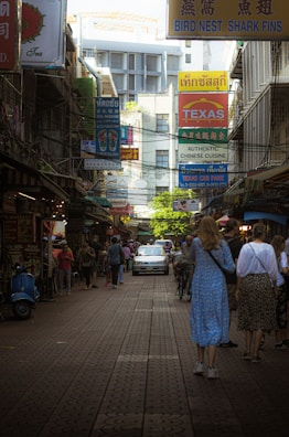 A bustling street scene lined with various colorful signs, including those advertising Chinese cuisine and bird nest products. People are walking along the street, and a car is driving down the center. The area has a crowded and lively atmosphere, with a mix of locals and tourists.