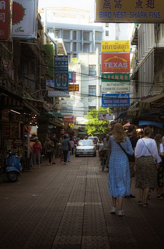 A bustling street scene lined with various colorful signs, including those advertising Chinese cuisine and bird nest products. People are walking along the street, and a car is driving down the center. The area has a crowded and lively atmosphere, with a mix of locals and tourists.
