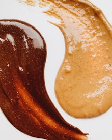 Swirls of creamy brown and light beige substances are spread on a smooth white surface, creating a visually appealing contrast. The texture appears glossy and rich, with the darker substance having a slightly darker, chocolate-like hue, while the lighter one resembles caramel or peanut butter.