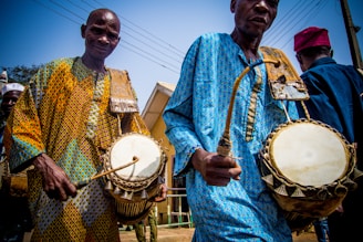 Two men are playing traditional drums while dressed in colorful, patterned clothing. The scene captures a vibrant and cultural performance, likely taking place outdoors as indicated by the visible wires and poles in the background.