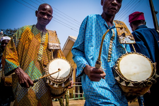 Two men are playing traditional drums while dressed in colorful, patterned clothing. The scene captures a vibrant and cultural performance, likely taking place outdoors as indicated by the visible wires and poles in the background.