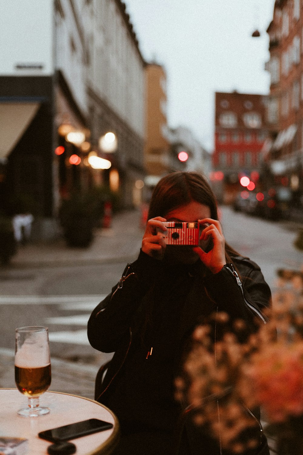 woman wearing black jacket using red and white point-and-shoot camera taking photo while sitting on chair