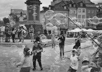 Children are playing with bubbles in a lively square surrounded by historic buildings, while onlookers watch and take photos. Large bubbles float through the air, adding to the joyful atmosphere.
