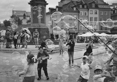 Children are playing with bubbles in a lively square surrounded by historic buildings, while onlookers watch and take photos. Large bubbles float through the air, adding to the joyful atmosphere.