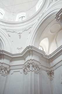 A highly detailed view of a building's interior architecture featuring grand, white, ornate arches and columns. There are intricately carved decorations and moldings at the tops of the columns and around the arches. Soft natural light filters through high windows, creating a serene and majestic atmosphere.