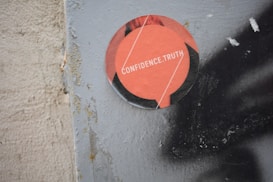 A circular sticker is attached to a weathered surface, featuring an orange design with the words 'CONFIDENCE.TRUTH' in white letters. The surface to which the sticker is adhered appears rough and textured, showing some signs of wear and rust.