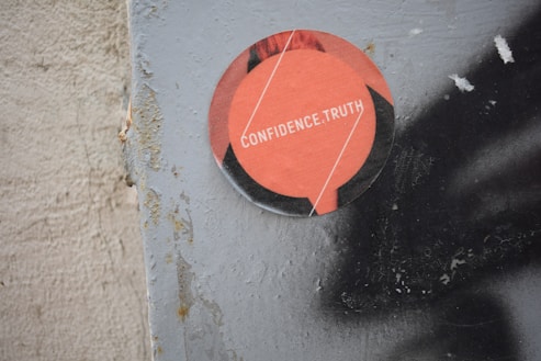 A circular sticker is attached to a weathered surface, featuring an orange design with the words 'CONFIDENCE.TRUTH' in white letters. The surface to which the sticker is adhered appears rough and textured, showing some signs of wear and rust.