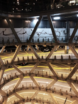 A multi-level spiral parking structure with concrete railings and visible yellow directional markings on the floors. Cars are parked on different levels, with some intricate wall art displayed above. The lighting is subdued with artificial lights illuminating the area.