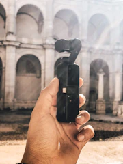 A hand holding a compact gimbal camera with a historic stone building in the background. The building features large arches and columns, and sunlight creates a warm tone with visible light rays.