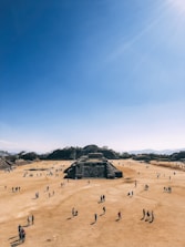 A large, open archaeological site with numerous people walking around. The central focus is a series of ancient stone structures and pyramids set against a clear blue sky. The landscape is dry and expansive, with hills in the background.