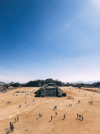 A large, open archaeological site with numerous people walking around. The central focus is a series of ancient stone structures and pyramids set against a clear blue sky. The landscape is dry and expansive, with hills in the background.