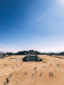 A large, open archaeological site with numerous people walking around. The central focus is a series of ancient stone structures and pyramids set against a clear blue sky. The landscape is dry and expansive, with hills in the background.