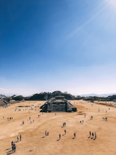 A large, open archaeological site with numerous people walking around. The central focus is a series of ancient stone structures and pyramids set against a clear blue sky. The landscape is dry and expansive, with hills in the background.