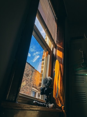 A cozy interior setting with a focus on a window showing a clear sky and adjacent brick wall. Inside, a camera mounted on a flexible gorillapod tripod sits beside a yellow cloth draped over a windowsill. Sunlight streams through the window, casting warm, soft light on the walls and objects.
