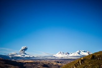 A snow-capped volcano is erupting under a clear blue sky, releasing a plume of smoke into the air. The landscape includes distant snowy mountains and a rocky, arid terrain in the foreground.