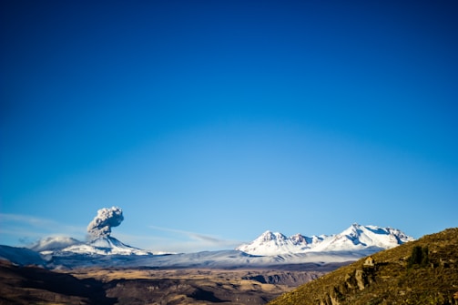A snow-capped volcano is erupting under a clear blue sky, releasing a plume of smoke into the air. The landscape includes distant snowy mountains and a rocky, arid terrain in the foreground.