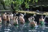 A group of people are participating in a ritual inside a pool of water, surrounded by stone carvings and greenery. They are facing a stone wall adorned with water spouts and offerings, creating a serene and spiritual atmosphere.