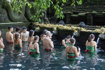 A group of people are participating in a ritual inside a pool of water, surrounded by stone carvings and greenery. They are facing a stone wall adorned with water spouts and offerings, creating a serene and spiritual atmosphere.