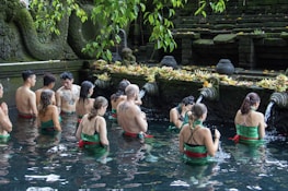 A group of people are participating in a ritual inside a pool of water, surrounded by stone carvings and greenery. They are facing a stone wall adorned with water spouts and offerings, creating a serene and spiritual atmosphere.