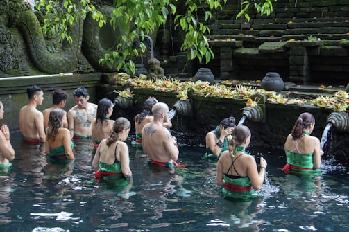 A group of people are participating in a ritual inside a pool of water, surrounded by stone carvings and greenery. They are facing a stone wall adorned with water spouts and offerings, creating a serene and spiritual atmosphere.