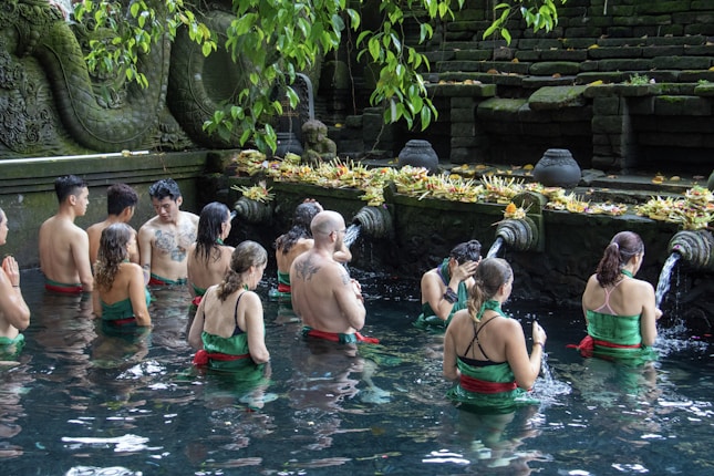 A group of people are participating in a ritual inside a pool of water, surrounded by stone carvings and greenery. They are facing a stone wall adorned with water spouts and offerings, creating a serene and spiritual atmosphere.