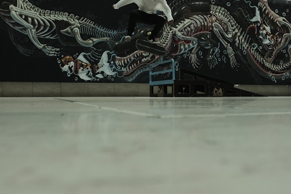 A skateboarder is in mid-air performing a trick above a ramp. The background features a detailed mural of a fantastical sea creature skeleton intertwining with abstract elements. The image is captured from a low angle, giving prominence to the mural and the motion of the skateboarder.